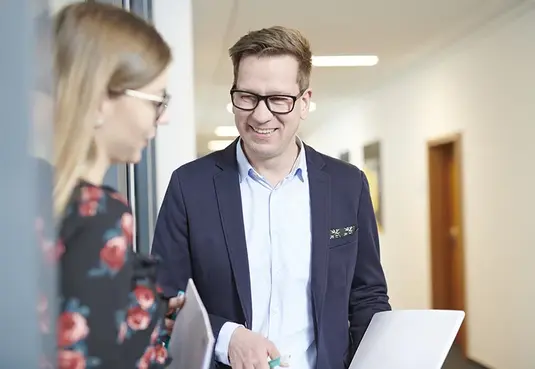 Bild mit zwei Personen mit Brille stehend in Business Kleidung auf einem Flur 